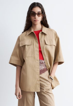 Femme aux longs cheveux foncés portant des lunettes de soleil marron et une chemise ample beige boutonnée sur un haut rouge et un pantalon beige, avec des bracelets au poignet.