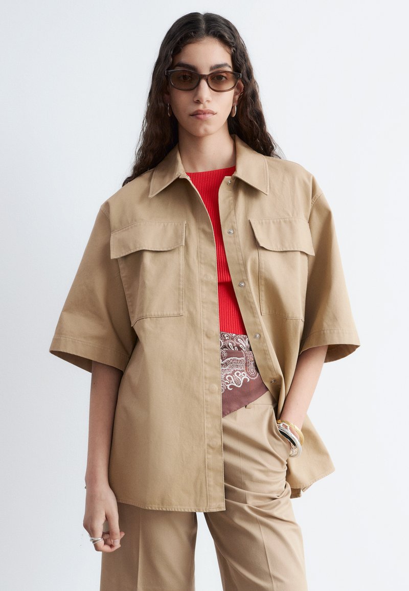 Femme aux longs cheveux foncés portant des lunettes de soleil marron et une chemise ample beige boutonnée sur un haut rouge et un pantalon beige, avec des bracelets au poignet.