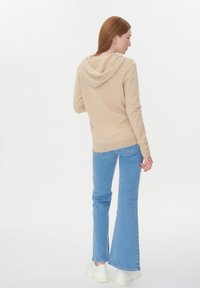 Rödhårig kvinna klädd i en beige luvtröja, ljusblå utsvängda jeans och vita sneakers, stående i en avslappnad pose med ryggen mot kameran.