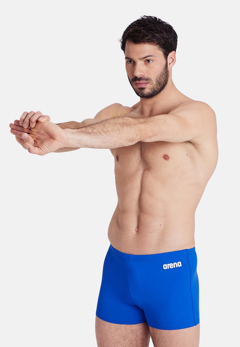 Bañador azul fabricado con tejido elástico, con un logo blanco en la cadera izquierda y un ajuste ceñido. Textura suave con detalles mínimos.