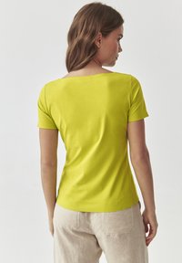 Limegrøn kortærmet skjorte med firkantet udskæring og glat tekstur, set bagfra, parret med lyse beige bukser.