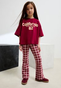Bordeauxfarbenes oversized T-Shirt mit weißem Schriftzug, kombiniert mit karierten Schlaghosen in Rottönen und Creme. Bordeauxfarbene Wildlederschuhe.