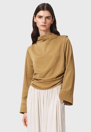 Vrouw met lang donker haar draagt een mosterdkleurige top met hoge hals en lange mouwen en een witte geplooide rok, staand tegen een egale achtergrond.