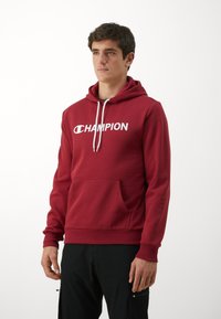 Roter Hoodie aus weichem Material mit einer Fronttasche, weißen Kordeln und einem großen weißen "CHAMPION"-Logo auf der Brust.