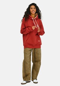 Červená mikina s kapucňou a predným vreckom, doplnená širokými nohavicami s leopardím vzorom. Biele tenisky a uvoľnená póza.