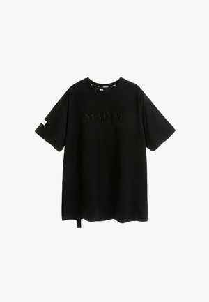 Camiseta negra oversized de manga corta. Presenta un logo bordado "MWM" en el frente y una correa lateral. Está hecha de algodón con una textura suave.