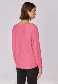 Maglione rosa a maniche lunghe con una vestibilità ampia, scollo a coste e texture morbida. Indossato con jeans scuri, mostrando una vista laterale.