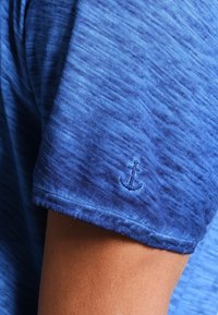 Niebieska koszula z krótkim rękawem, uszyta z teksturowanej tkaniny, z haftem kotwicy na rękawie. Rękawy są lekko podwinięte na brzegach.