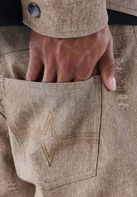 Tejido marrón texturizado con un bolsillo, que presenta un detalle de costura en forma de estrella en marrón más claro. Primer plano de una mano descansando en el bolsillo.