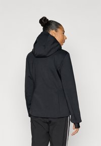 Čierna kapucňová bunda s priliehavým strihom, vyrobená z hladkého materiálu. Má vysoký golier a dlhé rukávy s otvorom na palec.