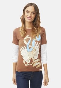 Camiseta corta de manga en color marrón superpuesta sobre una camisa de manga larga blanca, con un diseño gráfico de una figura alada y texto en la parte inferior.