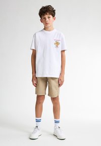 Witte katoenen t-shirt met een kleurrafisch "Key West", gecombineerd met beige shorts, witte sneakers en blauw gestreepte enkelsokken.