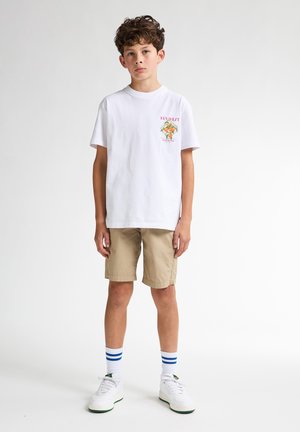 Camiseta de algodón blanca con un gráfico colorido de "Key West", combinada con pantalones cortos beige, zapatillas blancas y calcetines tobilleros a rayas azules.