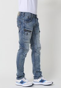 Pantalones cargo de mezclilla azul con múltiples bolsillos, costuras de contraste y un corte ajustado. Llevados con zapatillas blancas y azules.