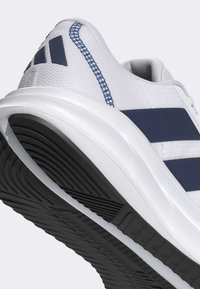 Chaussure de sport blanche avec des bandes bleu marine, semelle texturée et coutures bleues au talon, montrée en vue rapprochée sous un angle latéral et par le dessous.