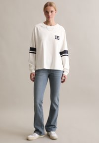 Frau trägt ein weißes Langarmshirt mit "15" und schwarzen Ärmelstreifen, hellblaue Jeans, weiße Sneakers und eine silberne Kettenhalskette.