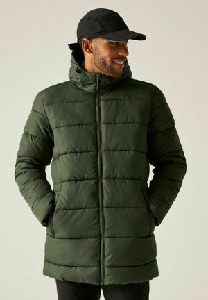 Mężczyzna w ciemnozielonej pikowanej kurtce na zamek i czarnej czapce, z rękami w kieszeniach, patrzący w bok i uśmiechający się.