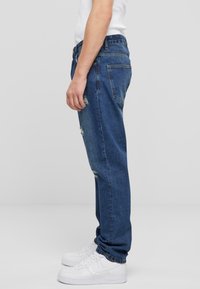 Jeans din denim albastru, cu croială lejeră, cu accente distruse, cinci buzunare și un design drept, asortați cu adidași albi.