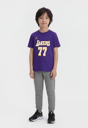 Fiatal fiú áll, lila Lakers #77-es pólót, szürke melegítőnadrágot és fekete Nike sportcipőt visel, egyszerű fehér háttér előtt.