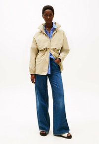 Giacca cropped color beige chiaro con zip frontale, vita stretta e maniche elastiche, indossata sopra una camicia a righe blu e jeans in denim a gamba larga.