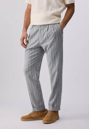 Uomo che indossa pantaloni grigi chiaro a righe sottili, camicia beige a maniche corte e sneakers casual beige con lacci, con una mano in tasca, in piedi.