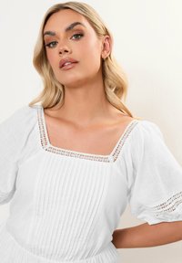 Vestido de lino blanco con escote cuadrado, bodice plisado y mangas abullonadas. Detalles de encaje en el escote y las mangas. Textura suave y ligera.