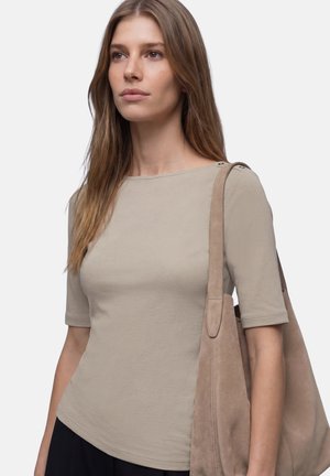 Kvinde med langt brunt hår iført en beige kortærmet top og bærende på en stor beige ruskind skuldertaske mod en ensfarvet baggrund.