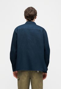 Άνδρας που φοράει μακρυμάνικο πουκάμισο σκούρου μπλε χρώματος και ελαι πράσινο παντελόνι cargo, στέκεται γυρισμένος με την πλάτη προς ένα απλό λευκό φόντο.