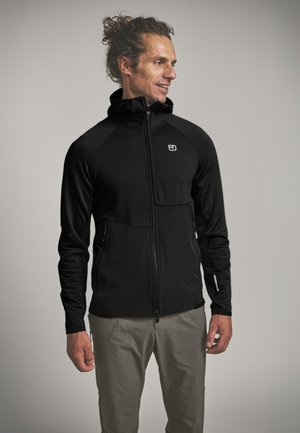 Mann mit lockigem Haar, der eine schwarze Kapuzenjacke mit Reißverschluss und graue Hosen trägt, steht und schaut seitlich vor einem neutralen Hintergrund.