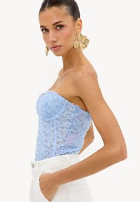 Femme aux cheveux lissés en arrière portant de grandes boucles d’oreilles fleurs dorées, un haut en dentelle bleu sans bretelles et un pantalon blanc taille haute avec les mains dans les poches.