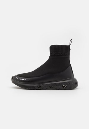 Bota preta de tornozelo com parte superior em malha texturizada, apresentando uma bainha canelada e uma sola de borracha grossa com padrões geométricos e detalhe de logotipo.