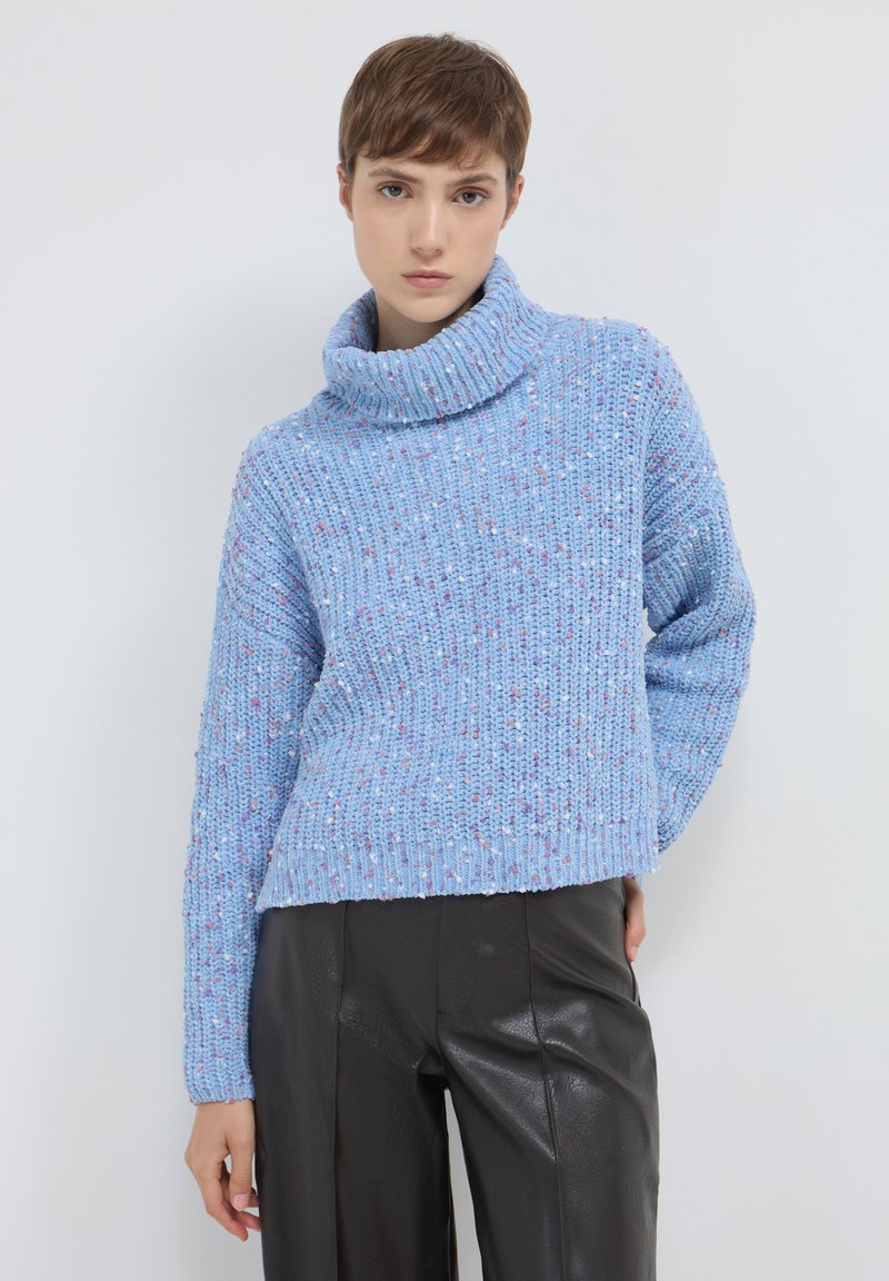 Persona che indossa un maglione blu a collo alto spesso e pantaloni in pelle neri, in piedi contro uno sfondo chiaro e uniforme con una mano dietro.