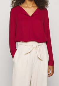Blusa rossa con scollatura a V, maniche lunghe e vestibilità ampia, abbinata a pantaloni plissettati a vita alta color crema con cintura annodata.