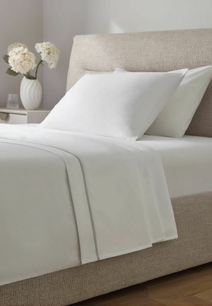 Beiges gepolstertes Bett mit weißen Kissen und weißer Bettwäsche mit dezenten grauen Linien, neben einem Nachttisch mit einer weißen Vase mit weißen Blumen.