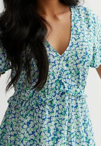 V-kaula-aukollinen kukkakuvioinen mekko sinisen, vihreän ja valkoisen sävyissä, jossa on röyhelöitä vyötäröllä ja hihoissa. Kevyt kangas ja liehuva leikkaus.