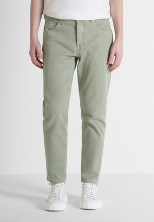 Man draagt lichtgroene slim-fit jeans en witte sneakers, staand tegen een effen achtergrond.