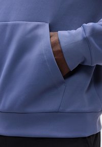 Main à la peau foncée insérée dans la poche avant d'un sweat à capuche bleu clair porté sur un pantalon noir.