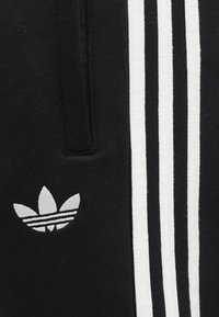 Čierne športové nohavice s bielym trojprúžkovým vzorom. Na prednej strane je umiestnené malé šedé vyšívané logo v tvare listu.