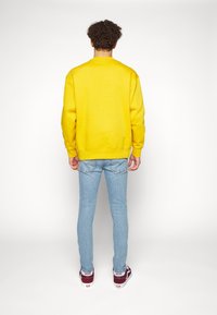 Gelbes Sweatshirt mit Rundhalsausschnitt, gerippten Bündchen und Saum, kombiniert mit hellblauen Slim-Fit-Jeans und roten Vans-Sneakern.