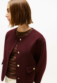 Bordeaux wollen blend jasje met gouden knopen, ronde halslijn en twee voorzakken. Gelaagd over een bruin gestreept shirt.