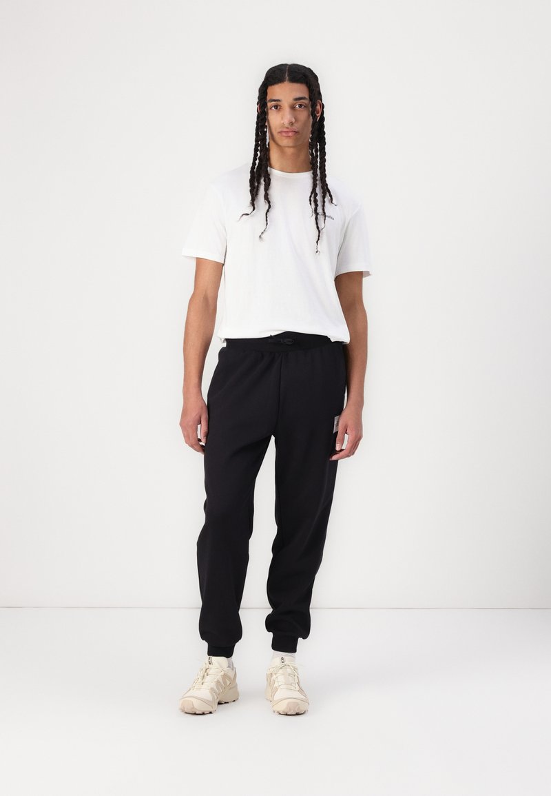 T-shirt en coton blanc associé à un pantalon de jogging noir avec une taille élastique et une poche latérale ; des baskets couleur crème complètent le look.