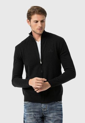 Schwarzer Reißverschluss-Pullover mit hohem Kragen, gerippten Bündchen und Saum, aus einem gestrickten Stoff gefertigt. Über einem weißen T-Shirt getragen.