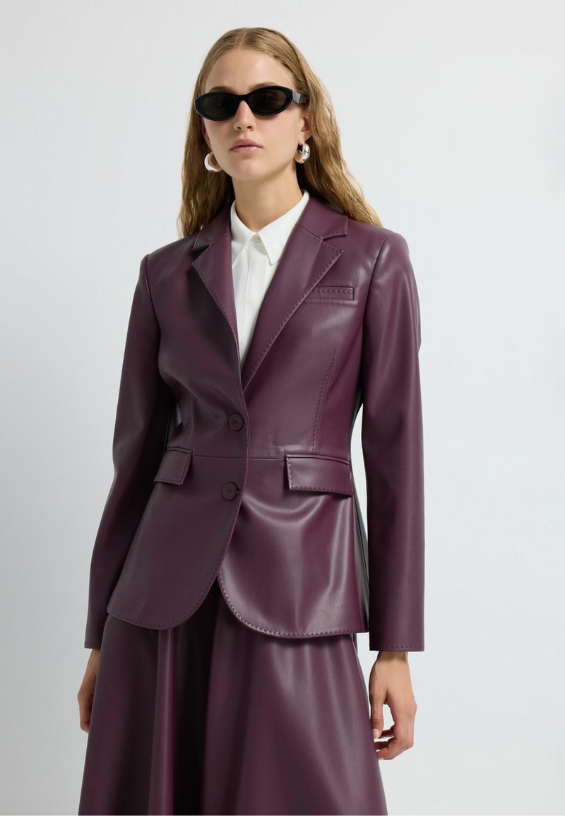 Blazer en cuir couleur prune avec deux boutons, revers crantés et poches latérales, porté sur une chemise blanche à col. Texture lisse et coupe ajustée.