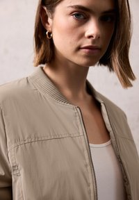 Veste bomber beige clair avec col côtelé et fermeture éclair, dotée d'une texture lisse et de détails de couture minimalistes, portée sur un haut blanc.