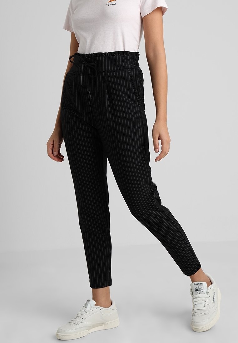Femme portant un pantalon noir à fines rayures fuselé avec taille à cordon, un t-shirt blanc graphique et des baskets blanches, debout devant un fond uni.