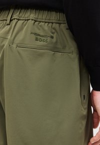 Ελιά πράσινο παντελόνι με ελαστική μέση διαθέτει ανάγλυφο λογότυπο "Porsche BOSS" πάνω από την πίσω τσέπη, φορεμένο με μαύρο τοπ και ορατό χέρι.