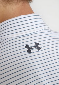 Valkoinen polo paita, jossa on sinisiä vaakasuoria raitoja, mustalla Under Armour -logolla takakauluksessa. Valmistettu kevyestä kankaasta.
