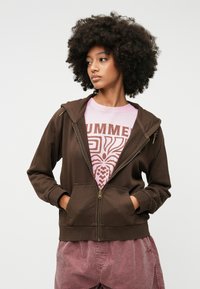 Καφέ φούτερ με φερμουάρ και κουκούλα, με μπροστινές τσέπες, ριμπ μοντελάκια, φορεμένο πάνω από ένα ροζ μπλουζάκι με γραφικό σχέδιο και μοτίβο.