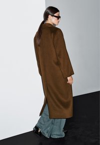 Brązowy wełniany płaszcz o długim kroju i bocznych rozcięciach, noszony na jasnoniebieskie dżinsy typu wide-leg. Czarne buty na szpilkach dopełniają stylizację.