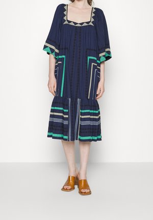 Kobieta w granatowej sukience midi z geometrycznymi wzorami w kolorach zielonym, beżowym i białym, w zestawieniu z beżowymi sandałami na słupku z odkrytymi palcami.